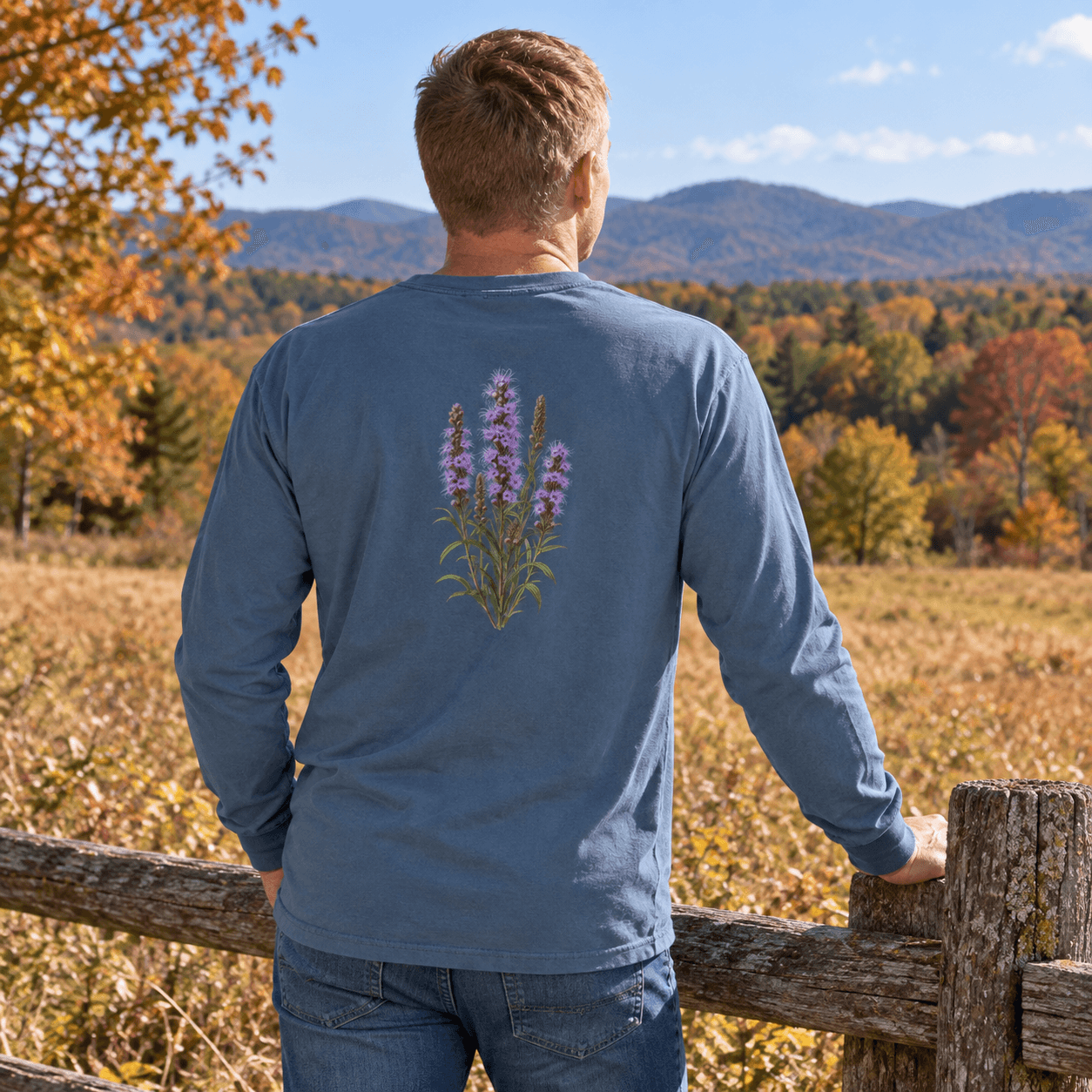 Rear view of a man in a blue long-sleeve shirt with a purple botanical print, beside a fence overlooking wooded hills. AI-generated promotional image.
